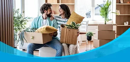 A smiling couple sits among moving boxes and houseplants in their bright new living room.